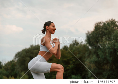 Doing squats. Young caucasian woman with slim body shape is in the fitness clothes outdoors 126314797