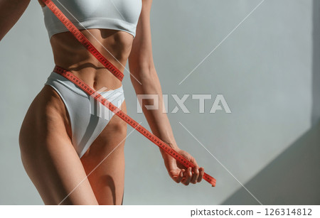 View of the waist and measuring tape in hands. Young caucasian woman with slim body shape is indoors in the studio View of the waist and measuring tape in hands. Young caucasian woman with slim body shape is indoors in the studio 126314812