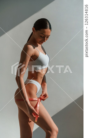 Using the measuring tape. Young caucasian woman with slim body shape is indoors in the studio Using the measuring tape. Young caucasian woman with slim body shape is indoors in the studio 126314816