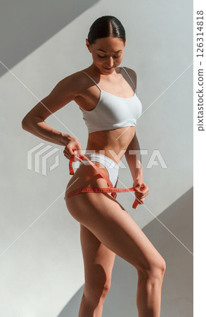 Using the measuring tape. Young caucasian woman with slim body shape is indoors in the studio 126314818