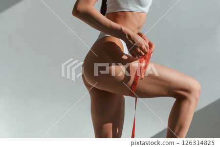 Using measuring tape on legs. Young caucasian woman with slim body shape is indoors in the studio 126314825