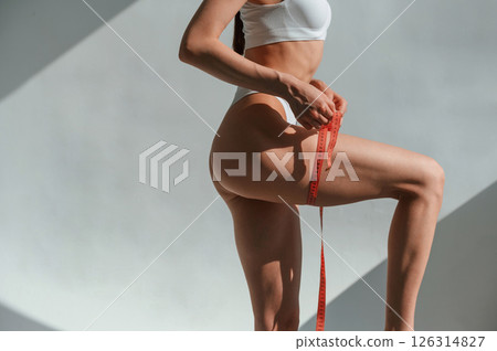 Using measuring tape on legs. Young caucasian woman with slim body shape is indoors in the studio 126314827