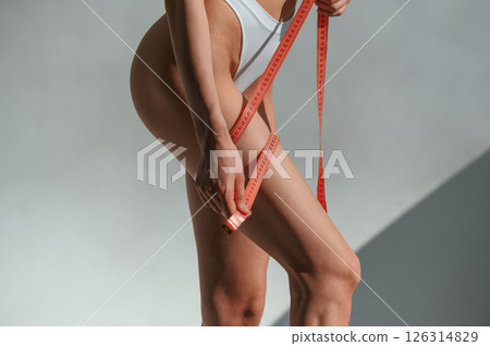 Using measuring tape on legs. Young caucasian woman with slim body shape is indoors in the studio 126314829