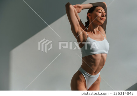 Indoors against the wall, beautiful sunlight. Young caucasian woman with slim body shape is indoors in the studio Indoors against the wall, beautiful sunlight. Young caucasian woman with slim body shape is indoors in the studio 126314842