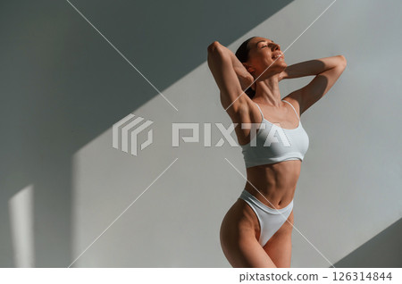 Indoors against the wall, beautiful sunlight. Young caucasian woman with slim body shape is indoors in the studio 126314844