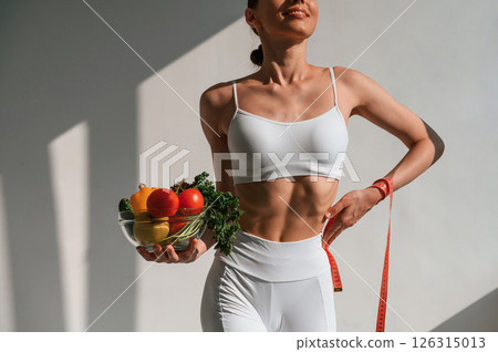 Standing against wall and holding healthy food. Young caucasian woman with slim body shape is indoors in the studio Standing against wall and holding healthy food. Young caucasian woman with slim body shape is indoors in the studio 126315013