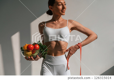 Standing against wall and holding healthy food. Young caucasian woman with slim body shape is indoors in the studio Standing against wall and holding healthy food. Young caucasian woman with slim body shape is indoors in the studio 126315014