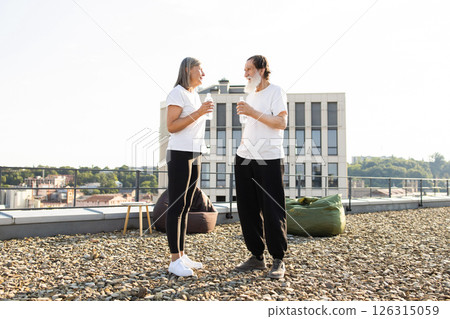 Senior man and woman standing on urban rooftop drinking bottled water, wearing casual clothing outdoors under sunlight Senior man and woman standing on urban rooftop drinking bottled water, wearing casual clothing outdoors under sunlight 126315059