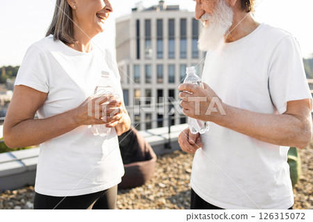 Senior man and woman exercising outdoors, standing rooftop, holding bottled water, enjoying discussion, wearing casual fitness outfits, showcasing active lifestyle and friendship 126315072