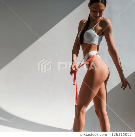 Red colored measuring tape in hands. Young caucasian woman with slim body shape is indoors in the studio 126315092