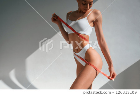 Measuring tape in hands. Young caucasian woman with slim body shape is indoors in the studio Measuring tape in hands. Young caucasian woman with slim body shape is indoors in the studio 126315109