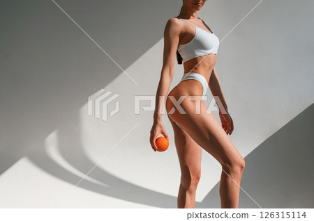 With an orange in hand. Young caucasian woman with slim body shape is indoors in the studio 126315114