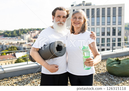 Senior man and woman outdoors on rooftop holding yoga mat and water bottle smiling showing active lifestyle and happiness. Captures healthy living, exercise, and connection against urban cityscape Senior man and woman outdoors on rooftop holding yoga mat and water bottle smiling showing active lifestyle and happiness. Captures healthy living, exercise, and connection against urban cityscape 126315118