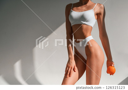 Standing, posing and holding an orange in hand. Young caucasian woman with slim body shape is indoors in the studio 126315123