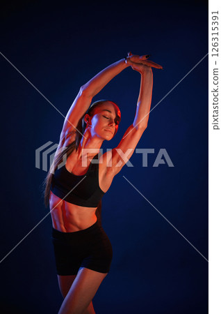 Stretching the hands. Beautiful muscular woman is indoors in the studio with neon lighting Stretching the hands. Beautiful muscular woman is indoors in the studio with neon lighting 126315391