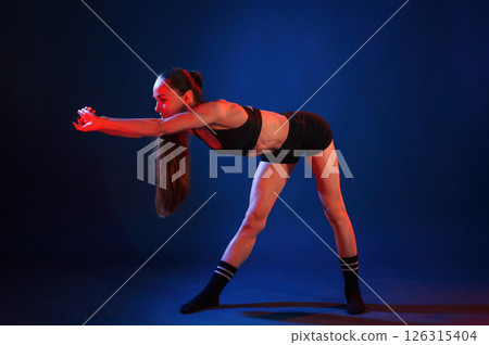 Doing sportive exerises. Beautiful muscular woman is indoors in the studio with neon lighting 126315404