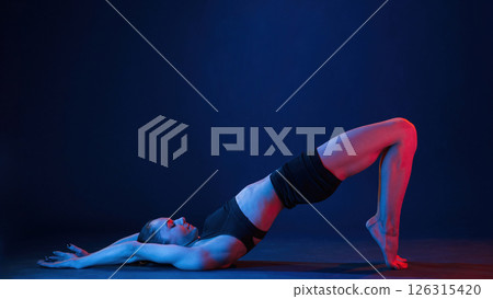 View from the side, lying down on the floor. Beautiful muscular woman is indoors in the studio with neon lighting View from the side, lying down on the floor. Beautiful muscular woman is indoors in the studio with neon lighting 126315420
