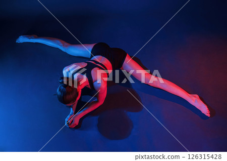 Performing the twine exercise. Beautiful muscular woman is indoors in the studio with neon lighting Performing the twine exercise. Beautiful muscular woman is indoors in the studio with neon lighting 126315428
