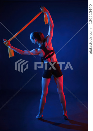 Yellow colored tape for stretching the hands. Beautiful muscular woman is indoors in the studio with neon lighting Yellow colored tape for stretching the hands. Beautiful muscular woman is indoors in the studio with neon lighting 126315440