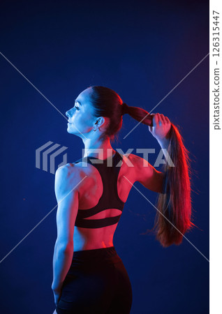Holding her hair, standing. Beautiful muscular woman is indoors in the studio with neon lighting 126315447