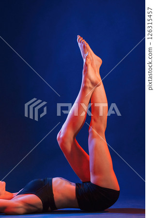 Lying down on floor and raising the legs up. Beautiful muscular woman is indoors in the studio with neon lighting Lying down on floor and raising the legs up. Beautiful muscular woman is indoors in the studio with neon lighting 126315457