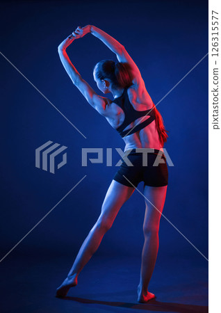 Back view. Warm up stretching exercises. Beautiful muscular woman is indoors in the studio with neon lighting Back view. Warm up stretching exercises. Beautiful muscular woman is indoors in the studio with neon lighting 126315577