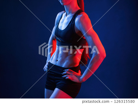 Standing with the hands on waist. Beautiful muscular woman is indoors in the studio with neon lighting Standing with the hands on waist. Beautiful muscular woman is indoors in the studio with neon lighting 126315580