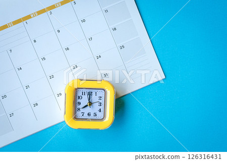 Calendar page and yellow clock on top of blue tabletop. 126316431
