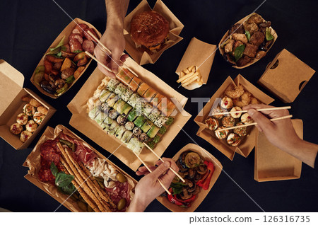 Top view of table with food on it, hands of the people taking it, eating. Meat, sushi, vegetables, French fries, burger, mushrooms Top view of table with food on it, hands of the people taking it, eating. Meat, sushi, vegetables, French fries, burger, mushrooms 126316735