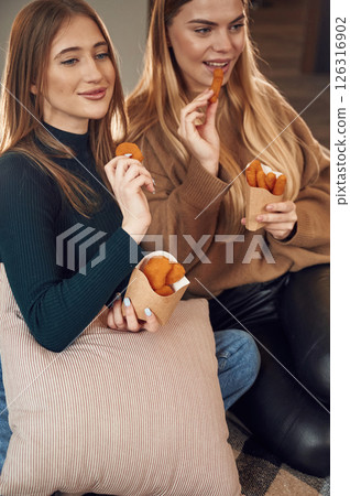Two women are sitting and eating chicken nuggets indoors together 126316902
