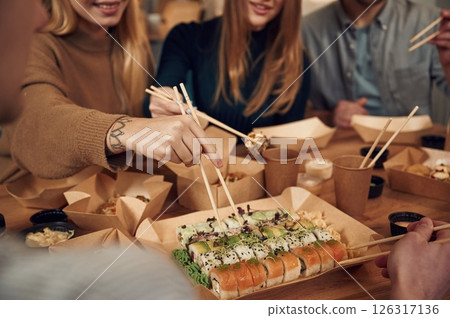 Using chopsticks. Friends are sitting indoors at home and eating sushi together Using chopsticks. Friends are sitting indoors at home and eating sushi together 126317136