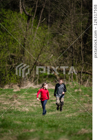 Two children joyfully run through a grassy field, surrounded by trees. 126317508