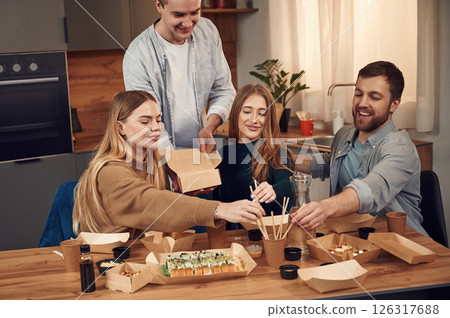 Man is standing. Friends are sitting indoors at home and eating sushi together Man is standing. Friends are sitting indoors at home and eating sushi together 126317688