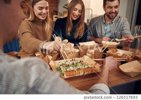 Using chopsticks. Friends are sitting indoors at home and eating sushi together Using chopsticks. Friends are sitting indoors at home and eating sushi together 126317693