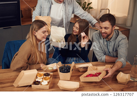 Friends are sitting indoors at home eating macarons, desserts and blueberries together Friends are sitting indoors at home eating macarons, desserts and blueberries together 126317707