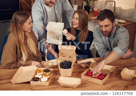Friends are sitting indoors at home eating macarons, desserts and blueberries together Friends are sitting indoors at home eating macarons, desserts and blueberries together 126317708