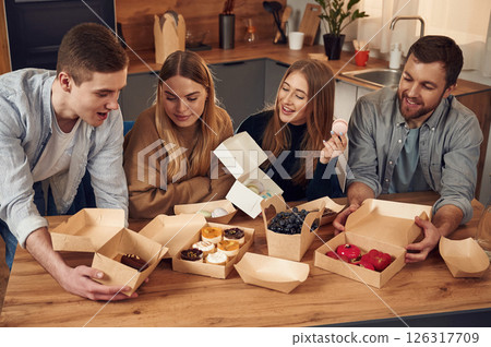 Friends are sitting indoors at home eating macarons, desserts and blueberries together Friends are sitting indoors at home eating macarons, desserts and blueberries together 126317709