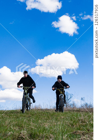 Two cyclists ascend a grassy hill under a vibrant blue sky dotted with fluffy white clouds. A day of outdoor adventure. 126317916