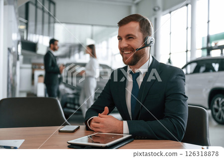 Man in headphones is sitting by table. Three people are working together in the car showroom Man in headphones is sitting by table. Three people are working together in the car showroom 126318878
