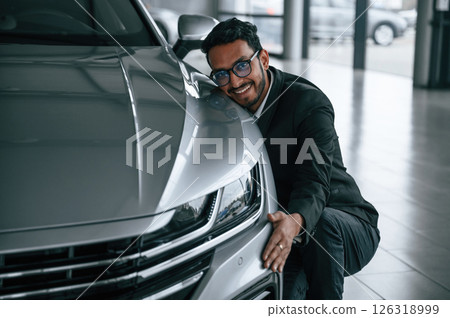 Embracing the transport. Handsome indian man in suit is in the car showroom with the vehicle indoors 126318999