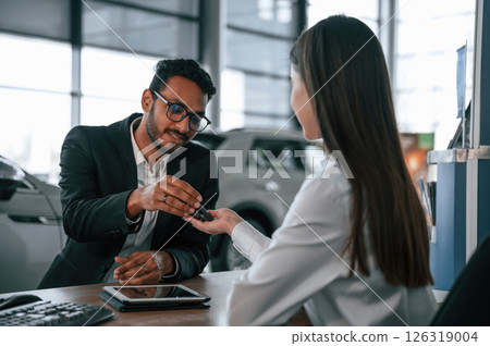 Woman is consulting man in the car showroom. Taking keys from the new automobile 126319004