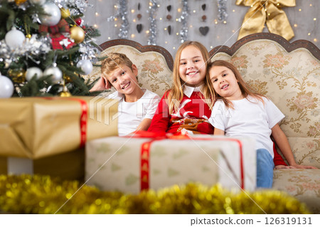 Satisfied children rejoice with New Year gifts, sitting on the sofa next to New Year tree Satisfied children rejoice with New Year gifts, sitting on the sofa next to New Year tree 126319131