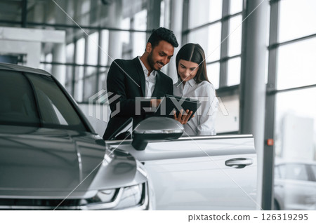Showing information in the graphic tablet. Man in formal clothes is consulting woman about the automobile in the car dealership 126319295