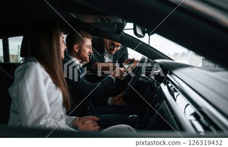 Side view. Car showroom worker is consulting man and woman that are sitting inside of automobile Side view. Car showroom worker is consulting man and woman that are sitting inside of automobile 126319432