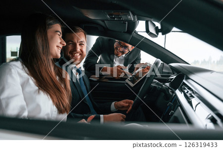 Side view. Car showroom worker is consulting man and woman that are sitting inside of automobile Side view. Car showroom worker is consulting man and woman that are sitting inside of automobile 126319433