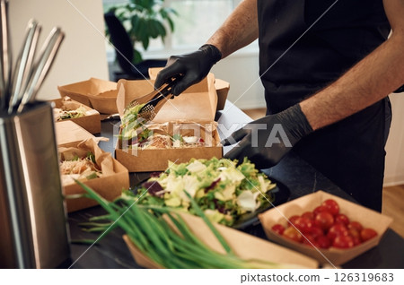 Adding vegetables to the burger. Man is packing food into the paper eco boxes. Indoors, restaurant Adding vegetables to the burger. Man is packing food into the paper eco boxes. Indoors, restaurant 126319683