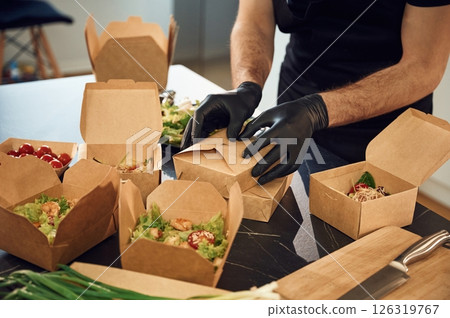In the black gloves. Man is packing food into the paper eco boxes. Indoors, restaurant 126319767