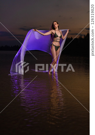 Young slender girl in a swimsuit posing in the water at sunset with purple colored piece of fabric 126319820