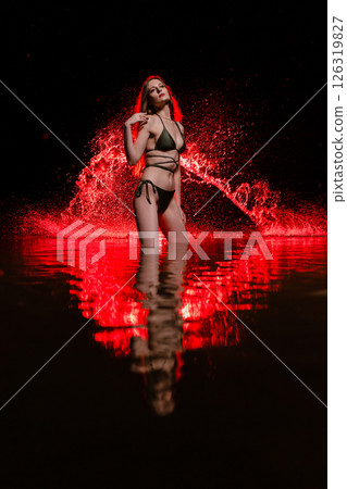 Young slender girl in a swimsuit posing in the water at sunset at the background of red colored water droplets flying in the air 126319827