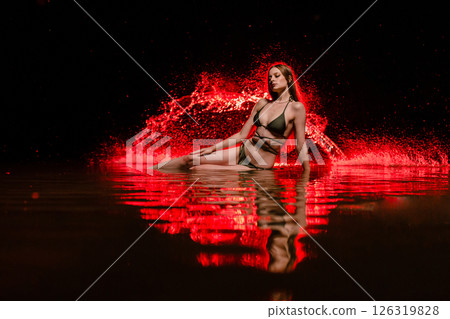 Young slender girl in a swimsuit posing in the water at sunset at the background of red colored water droplets flying in the air 126319828